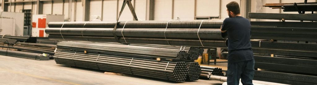 Steel pipes being maneuvered in an industrial facility by a worker using a crane.