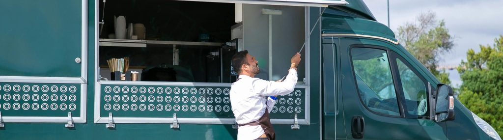 Side view young waiter in apron standing outside food truck and opening counter door in green lush park